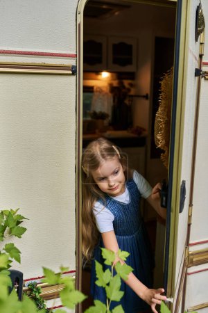 fröhliches Mädchen mit blonden Haaren im Jeanskleid, das aus dem modernen Wohnwagen schaut, glückliche Kindheit