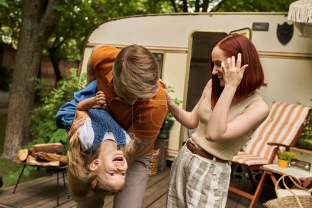 man playing with laughing daughter near happy wife and mobile home in trailer park, quality time