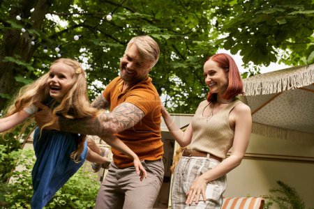 tattooed man playing with excited daughter near cheerful wife and trailer home, quality time
