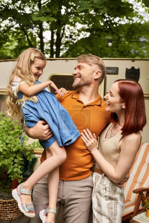 cheerful man holding cute daughter near smiling wife and trailer home on park, family bonding