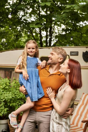 Téléchargez les photos : Homme souriant tenant fille mignonne près de femme heureuse et maison de remorque sur le parc, lien de famille - en image libre de droit