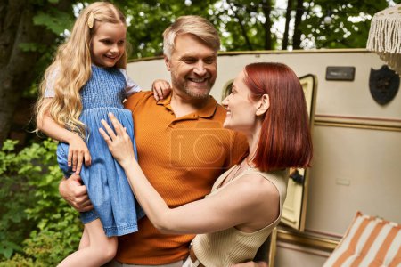 smiling man holding cute daughter near pleased wife and mobile home in trailer park, leisure