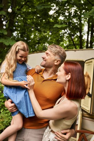 father holding laughing daughter near pleased wife and mobile home in trailer park, bonding