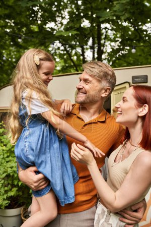 father holding cheerful daughter near pleased wife and modern trailer home, leisure and adventure