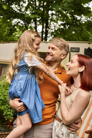 happy man holding adorable daughter near smiling wife and modern trailer home, leisure and adventure