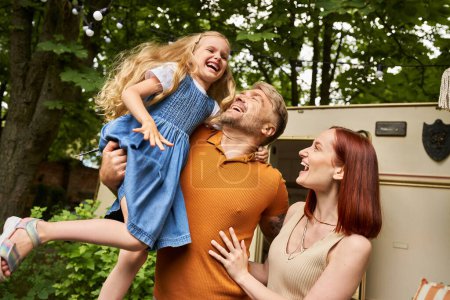 Téléchargez les photos : Père tenant fille ravie près souriante épouse et maison remorque moderne, amusant et rire - en image libre de droit