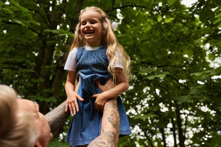 Photo for Overjoyed girl laughing in hands of tattooed father in green park, leisure and playing together - Royalty Free Image