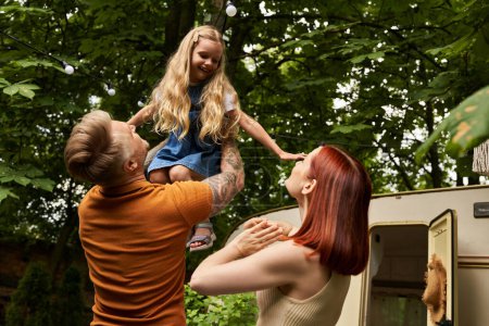 Foto de Hombre tatuado sosteniendo a la hija llena de alegría en las manos cerca de la esposa y el remolque moderno en casa, ocio familiar - Imagen libre de derechos