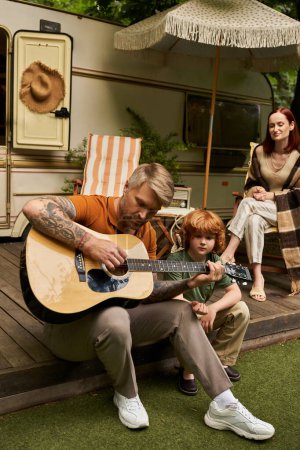 Tätowierter Mann spielt Akustikgitarre vor lächelndem Sohn in der Nähe von Familie und Wohnwagen, verbindende Momente