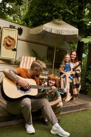 Tätowierter Mann spielt Akustikgitarre für glücklichen rothaarigen Sohn in der Nähe von Familie und modernem Wohnwagen
