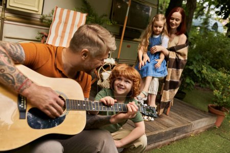 tattooed man playing acoustic guitar to cheerful redhead kid near family and modern trailer home