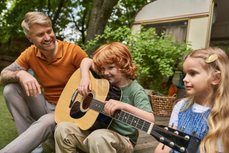 redhead boy playing guitar near smiling father and sister next to trailer home, creative activities