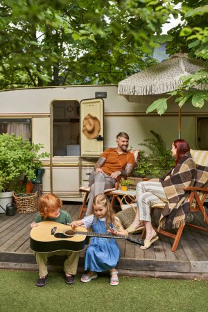 Téléchargez les photos : Enfants apprenant à jouer de la guitare acoustique tandis que les parents assis dans des chaises longues près de la maison de remorque - en image libre de droit