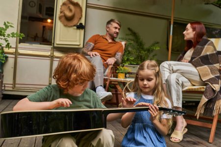 Téléchargez les photos : Frères et sœurs apprenant à jouer de la guitare tandis que les parents assis près de la maison de remorque, loisirs et plaisir - en image libre de droit