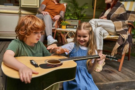 Téléchargez les photos : Rousse garçon apprendre à jouer de la guitare près joyeuse soeur et les parents assis à la maison de remorque - en image libre de droit