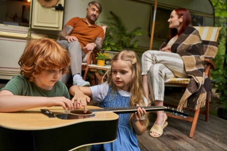 cute siblings learning to play guitar near parents sitting at talking next to trailer home
