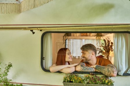 glücklich tätowierter Mann und rothaarige Frau, die sich im Fenster eines modernen Wohnwagens anschauen