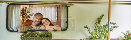 excited and stylish couple looking out window of mobile home around greenery in trailer park, banner