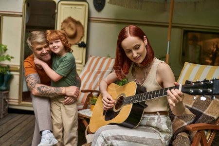 Téléchargez les photos : Femme souriante jouant de la guitare acoustique près de mari tatoué embrassant fils à côté de la maison de remorque - en image libre de droit