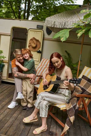 Téléchargez les photos : Femme joyeuse jouant de la guitare acoustique près de mari tatoué embrassant fils à côté de la maison de remorque - en image libre de droit