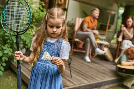 Téléchargez les photos : Fille tenant roquettes de badminton et navette près de la famille et la maison de remorque sur fond flou - en image libre de droit