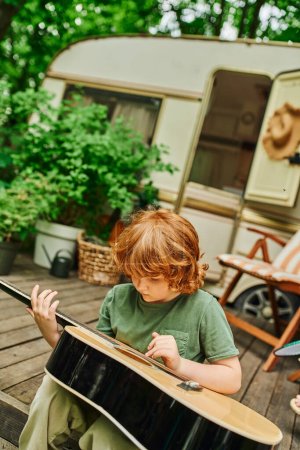 Téléchargez les photos : Rousse garçon apprendre à jouer de la guitare acoustique tout en étant assis près de la maison mobile dans le parc de remorques - en image libre de droit