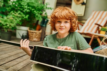 rothaariger Junge mit Akustikgitarre blickt in die Kamera, während er in der Nähe eines Wohnmobils auf einem Trailerpark sitzt