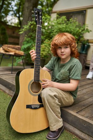 Foto de Confiado pelirrojo chico con la guitarra acústica mirando a la cámara mientras está sentado cerca de casa remolque moderno - Imagen libre de derechos