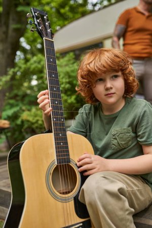 niño pelirrojo soñador con guitarra acústica mirando hacia otro lado mientras está sentado cerca de casa remolque moderno
