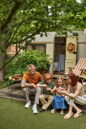Frau zeigt Kindern, die neben modernem Wohnwagen sitzen, wie man mit Seifenblasen spielt