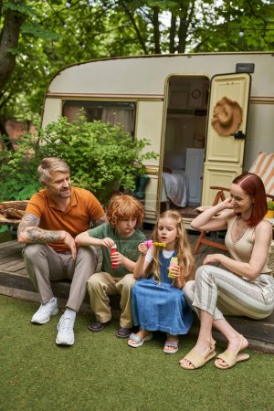 entzückende Kinder pusten Seifenblasen neben lächelnden Eltern neben modernem Wohnwagen, Freizeit und Spaß