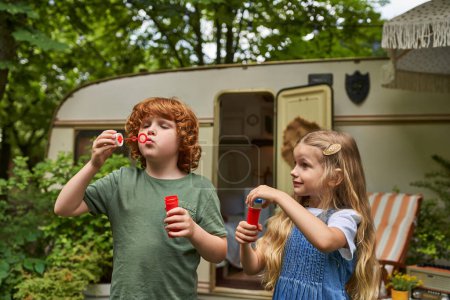 Photo for Redhead brother and blonde sister blowing soap bubbles near trailer home, siblings playing together - Royalty Free Image