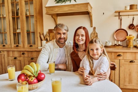 Foto de Padres felices con linda hija mirando a la cámara cerca de frutas frescas y jugo de naranja en la cocina - Imagen libre de derechos