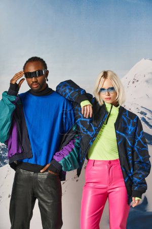 multiethnisches Paar in warmen, lebendigen Outfits mit Sonnenbrille vor Bergkulisse, Winterkonzept