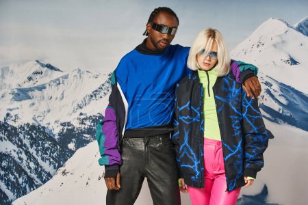 Téléchargez les photos : Jeune couple interracial étreignant et regardant caméra avec toile de fond de montagne, concept d'hiver - en image libre de droit