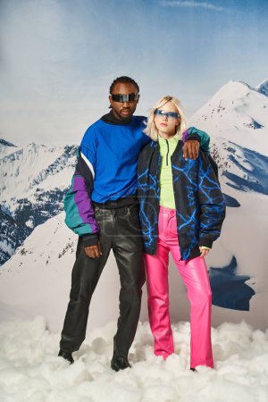 stylisches multiethnisches Paar, das sich umarmt und vor verschneiter Bergkulisse posiert, Winterkonzept