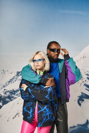modisches multiethnisches Paar im Winteroutfit mit stylischer Brille, die sich umarmt, Bergkulisse