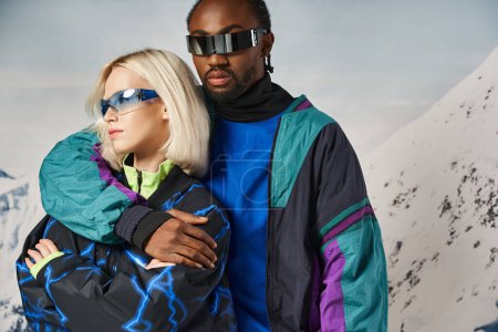 pareja elegante posando en traje cálido con gafas vibrantes con telón de fondo de montaña, concepto de invierno