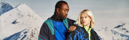 stylish multicultural couple in bold clothes with snowy mountain on backdrop, winter concept, banner