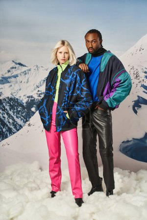 stylisches junges Paar in warmer Kleidung posiert mit Hand auf Schulter vor verschneiter Kulisse, Winterkonzept