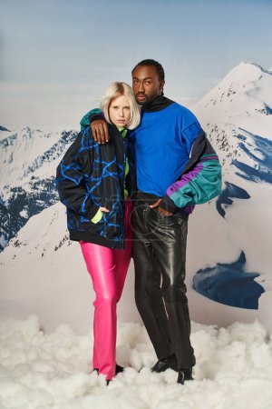 Modernes multiethnisches Paar in stilvoller warmer Kleidung, das sich umarmt und in die Kamera blickt, Winterkonzept