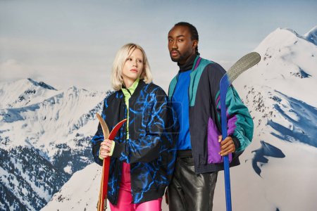 stylish multicultural couple in vibrant clothes posing with skis and hockey stick, winter concept