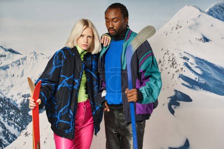 stylish multicultural couple posing with skis and hockey stick, hand on shoulder, winter concept