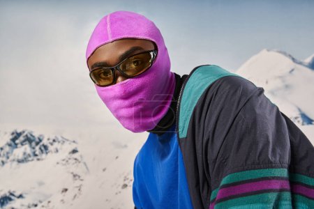 schöner stylischer Mann mit pinkfarbener Sturmhaube und Sonnenbrille vor verschneiter Kulisse, Winterkonzept