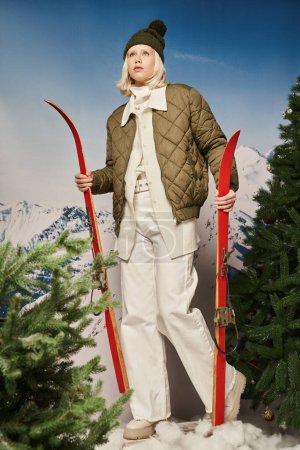 stylische blonde Frau mit Hut und Winterjacke mit Skiern neben Kiefern, Winterkonzept