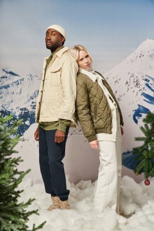 stylish multicultural couple in winter jackets posing back to back next to fit trees, winter concept