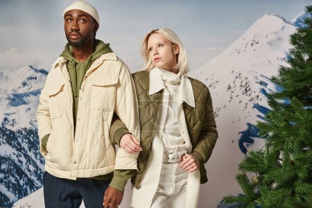 couple à la mode dans des vestes chaudes à côté de pin posant avec la main sur le bras, concept d'hiver