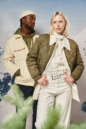 atractiva pareja con estilo en ropa de abrigo posando junto a abeto con telón de fondo de montaña, invierno