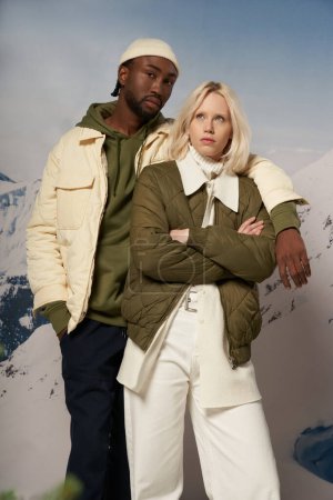 diverse fashionable couple in warm attire posing on snowy backdrop with mountain, winter concept