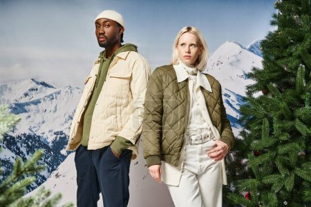 beautiful stylish couple wearing warm modish jackets and posing next to fir trees, winter concept